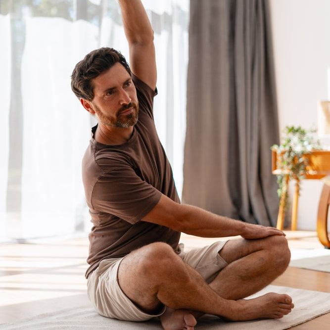 A man in a brown t-shirt and beige shorts sits cross-legged on a yoga mat in a bright room, stretching his arm overhead in a yoga pose.